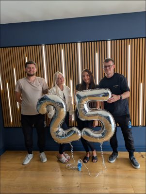 Four people stand indoors holding large silver balloons shaped like the number 25, celebrating an Everlast 25th anniversary and building legacy together.