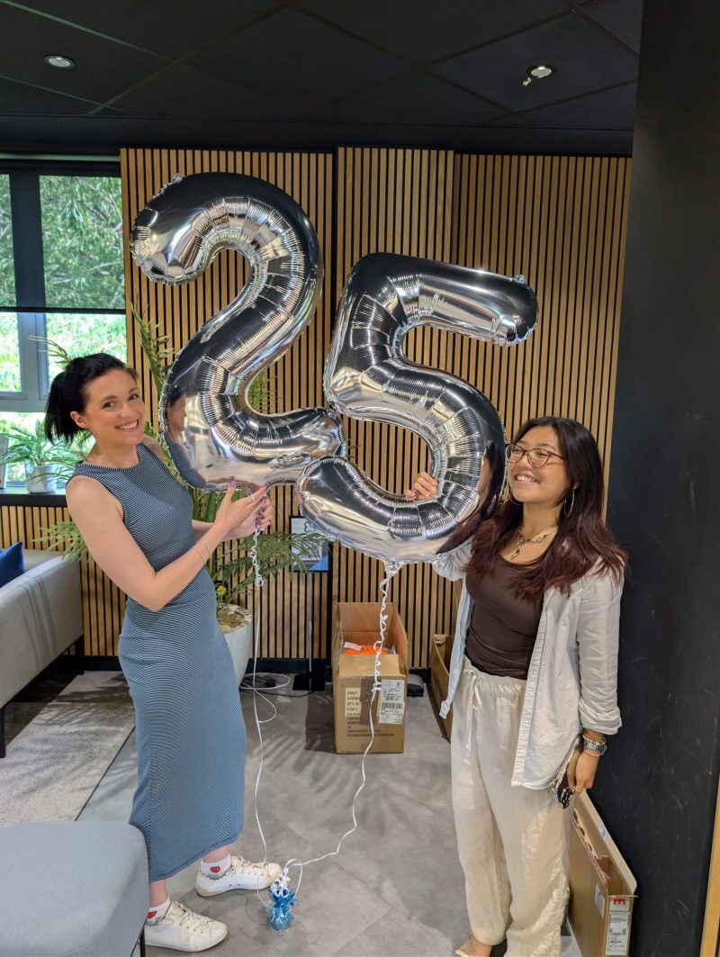 Two people smiling and holding large silver balloons shaped as the number 25.
