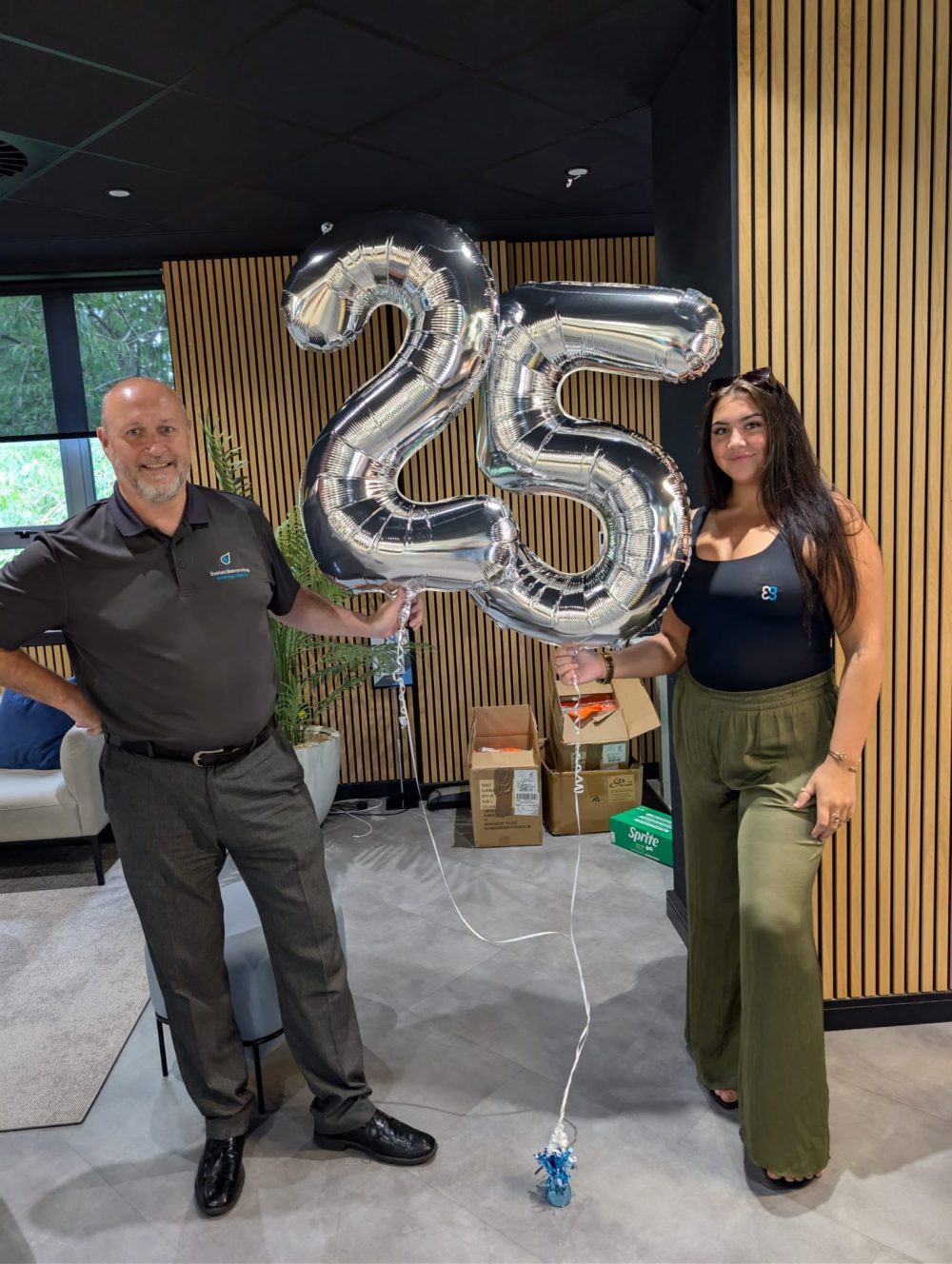 Two people holding silver balloons shaped as the number 25 in an office setting.