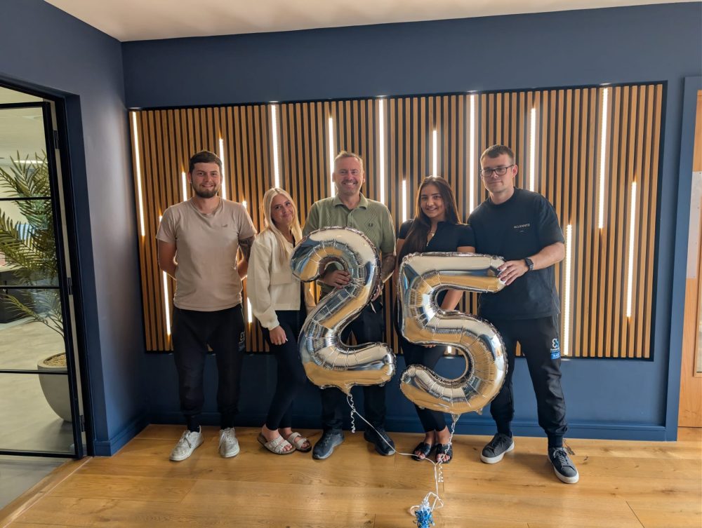 Five people smiling indoors, holding silver 25 balloons, standing on wooden floor.