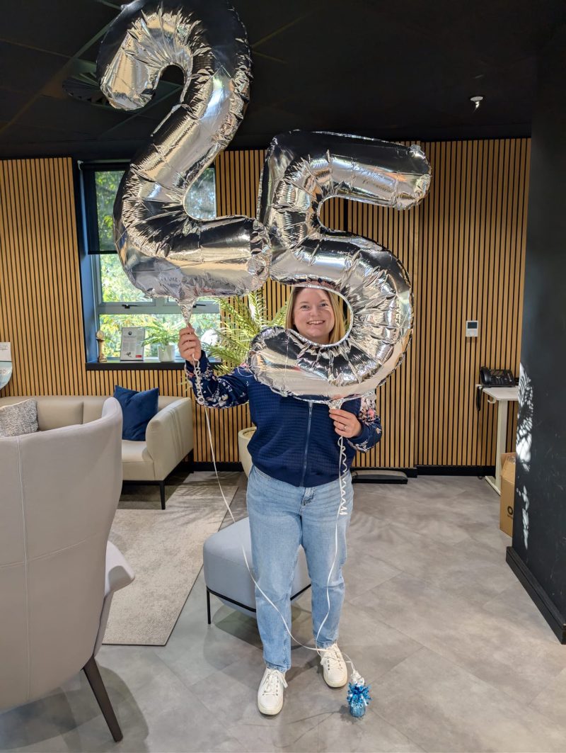 Woman holding large silver 25 balloons in a modern room, smiling at the camera.