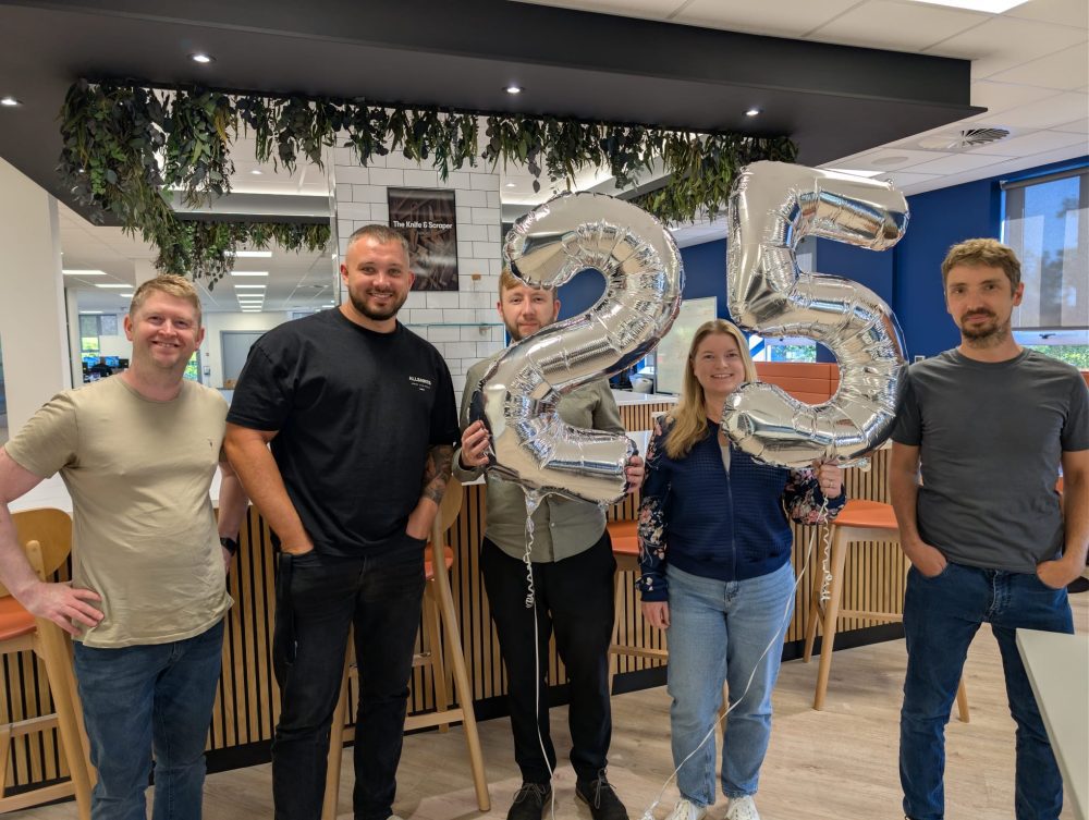 Image – 2025-08-15T084450.445 Five people standing indoors holding silver balloons shaped as the number 25.