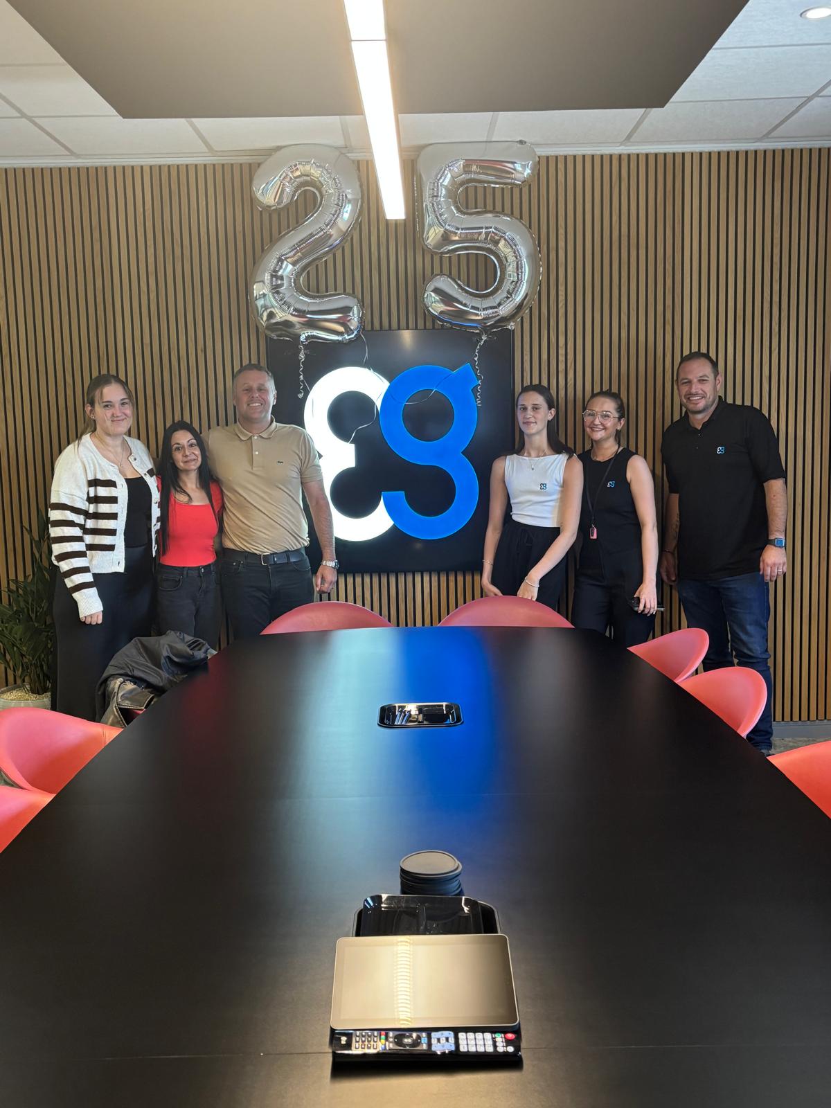 Six people stand in an office by 25 balloons and a blue and white logo on the wall.