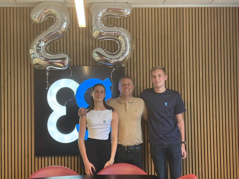 Three people pose indoors with silver 25 balloons and a blue-and-black wall sign behind them.