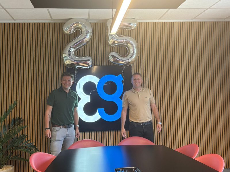 Two smiling men stand by silver 25 balloons and a glowing blue sign in an office.