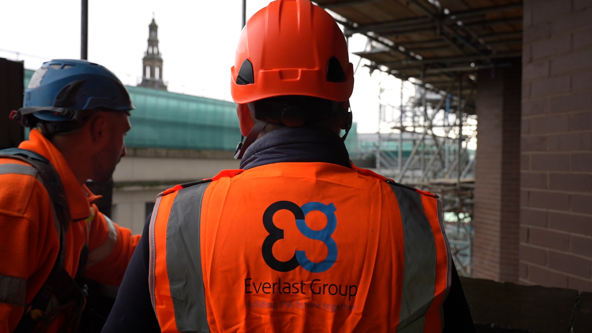 Two construction workers in orange vests and helmets stand at Leeds City Exchange site.