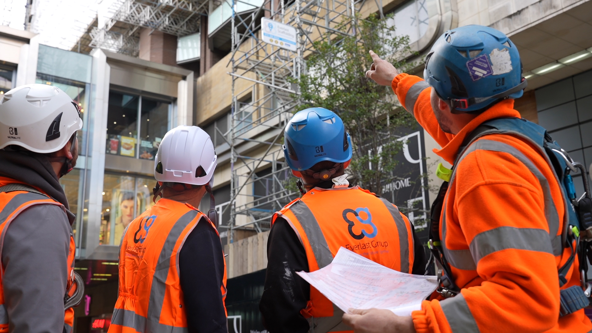 Four construction workers in safety gear discuss scaffolding outside Leeds City Exchange.