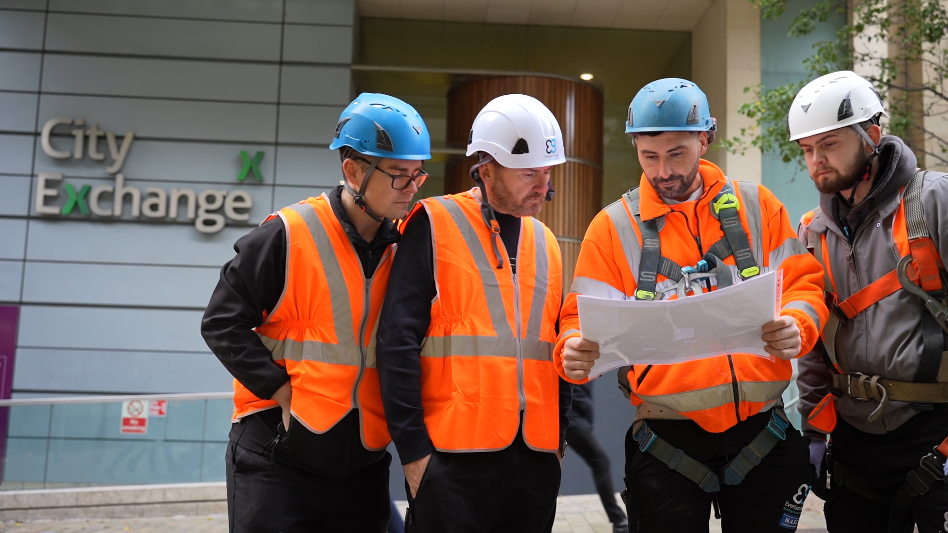 Four construction workers in orange vests review plans outside Leeds' City Exchange building.