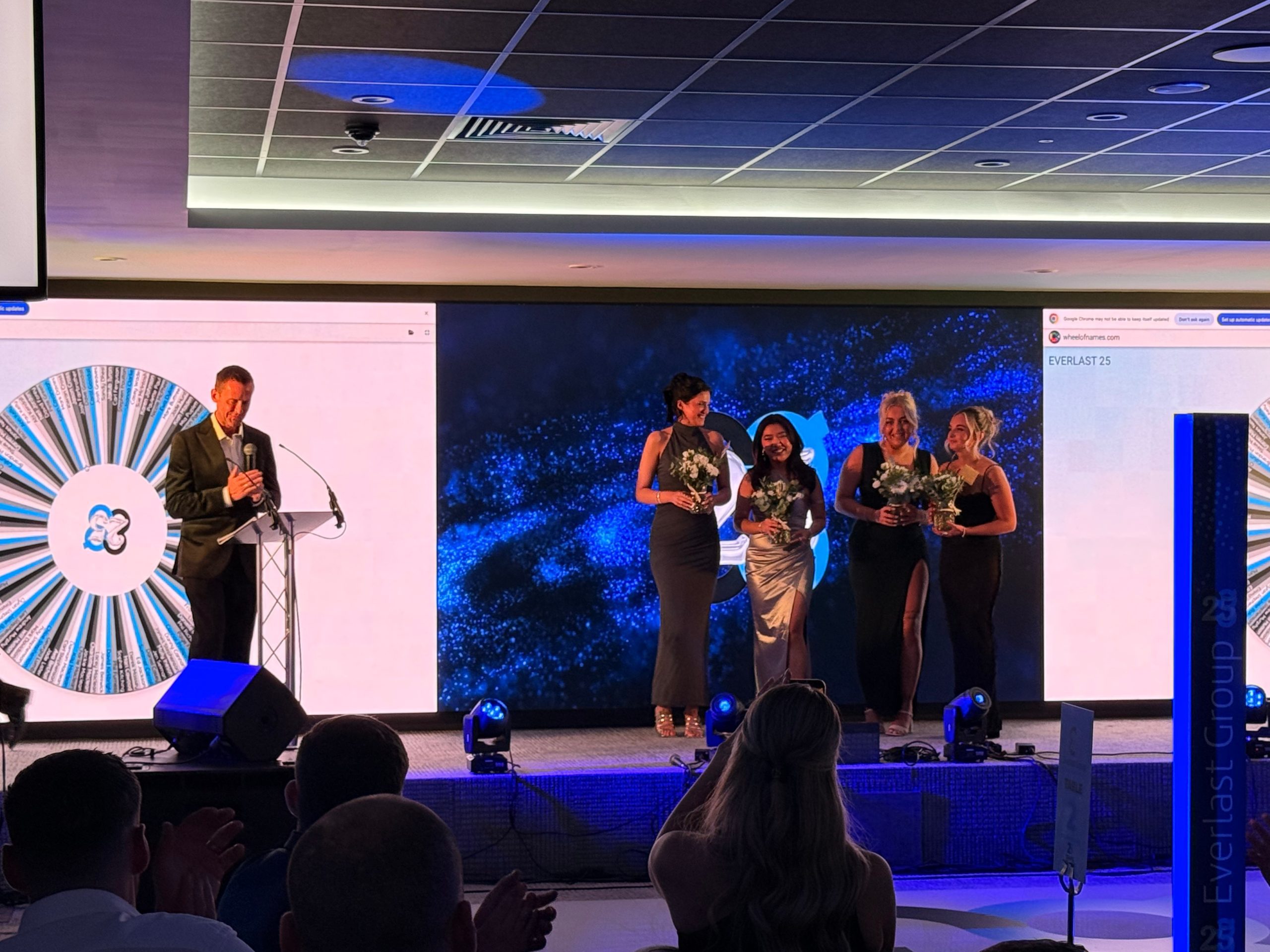 Five people on stage at the 25 Years Anniversary Event, with screens and lights in the background.