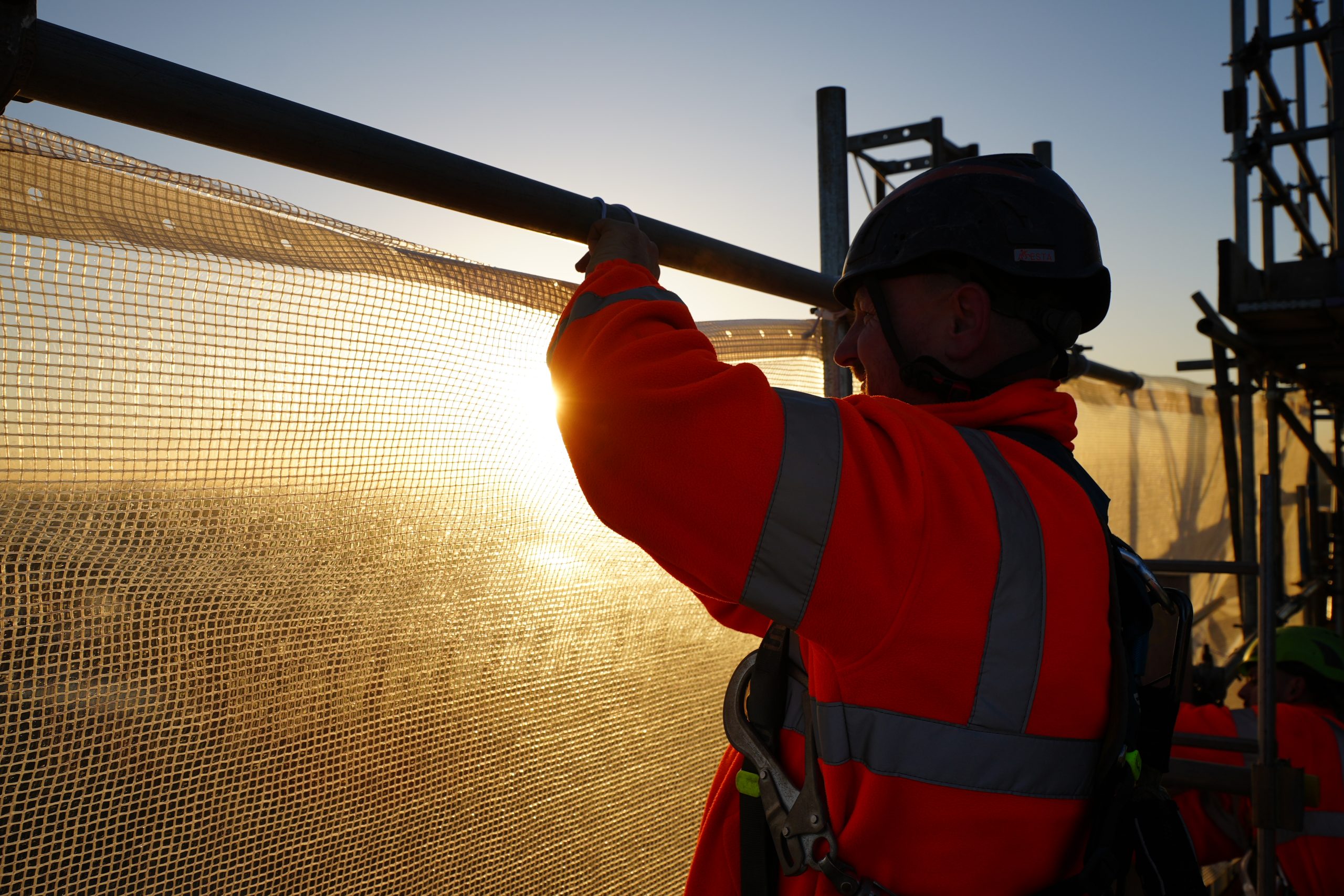 Construction worker in safety gear fitting mesh netting at City Exchange, Leeds, at sunset.