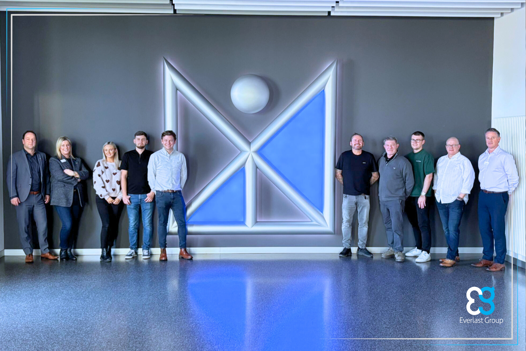 Ten people stand in front of a large blue and white Layher Germany geometric wall logo.