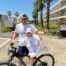 Man and child in matching shirts pose by a bike on a sunny path near palm trees and Leeds buildings.