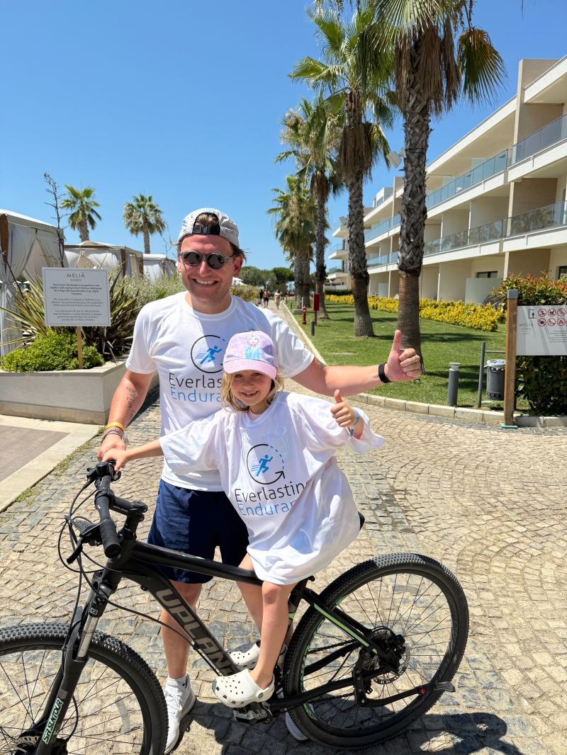 Man and child in matching shirts pose by a bike on a sunny path near palm trees and Leeds buildings.