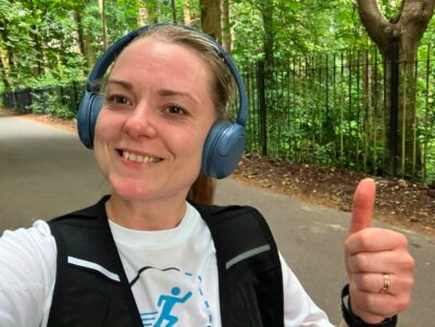 Woman wearing headphones gives a thumbs up whilst jogging in a green, wooded Leeds park.